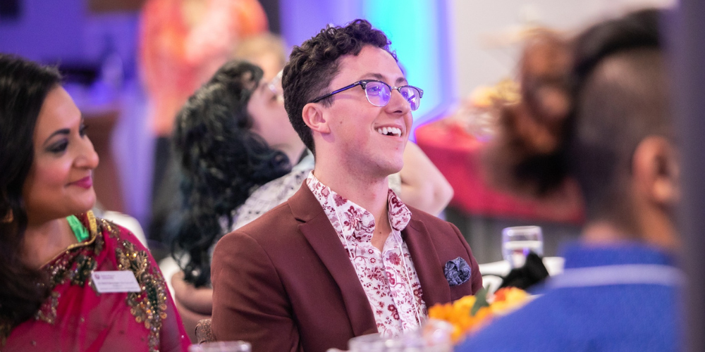 A person in a maroon suit sits at a table during an event