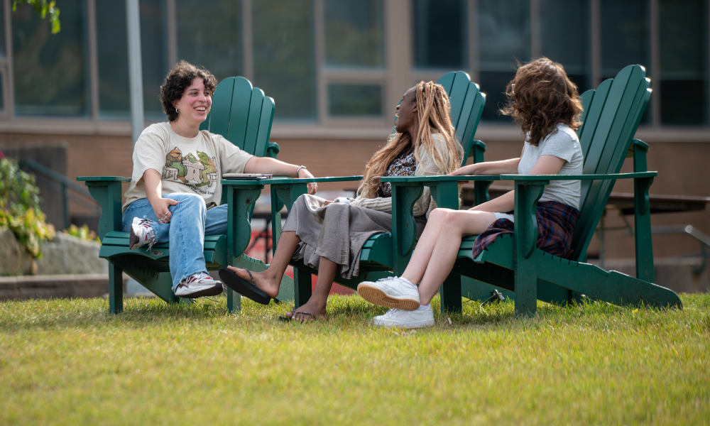 Three students sit on chairs outside on campus